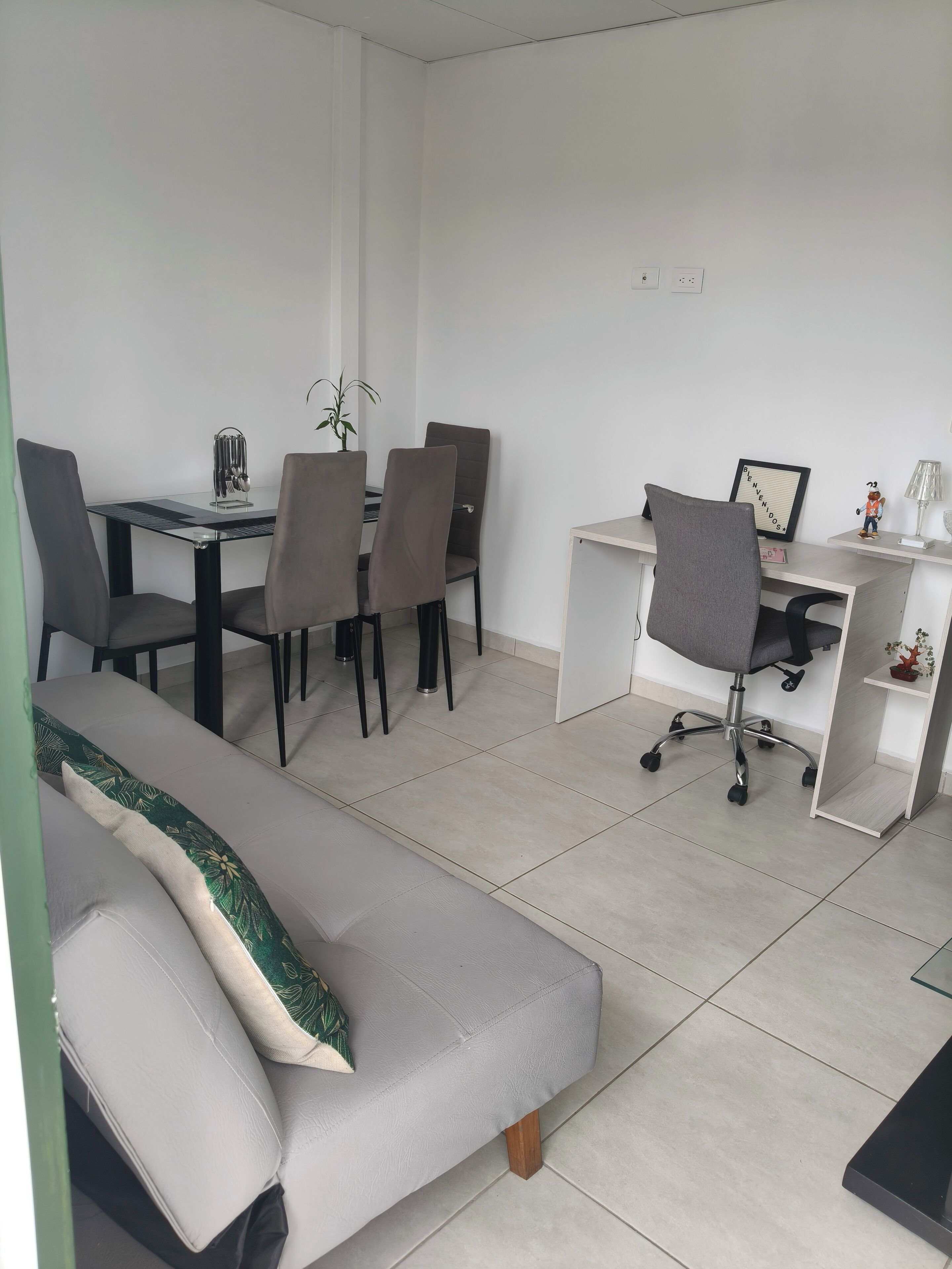 Een moderne, minimalistisch ingerichte kamer met een bank, een eettafel met stoelen, een bureau en een stoel, en een kleine plant.