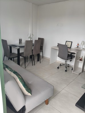 Et moderne, minimalistisk værelse med en sofa, et spisebord med stole, et skrivebord med stol og en lille plante.