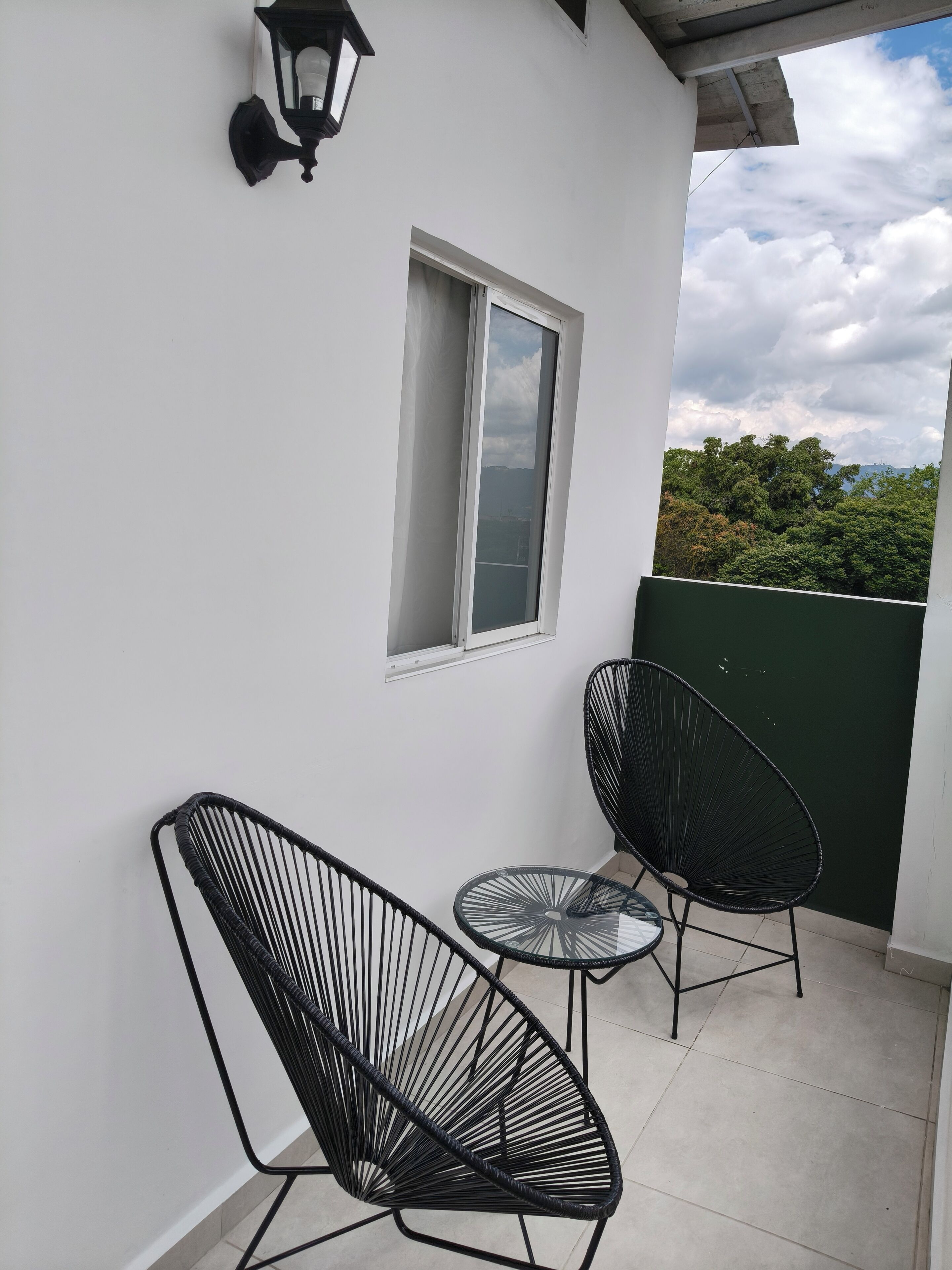 Un balcón con dos sillas de alambre negro, una mesita redonda y una lámpara de pared.