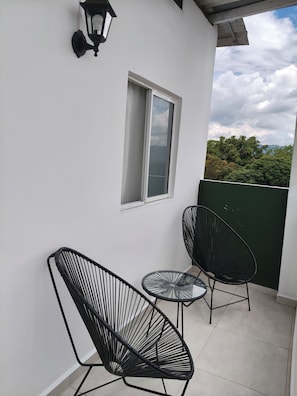 En balkon med to stole med sorte wirestel, et lille rundt bord og en vægmonteret lampe.