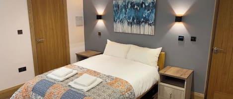A bedroom with a bed, two bedside tables, a nightstand, a wall-mounted painting, and two doors.