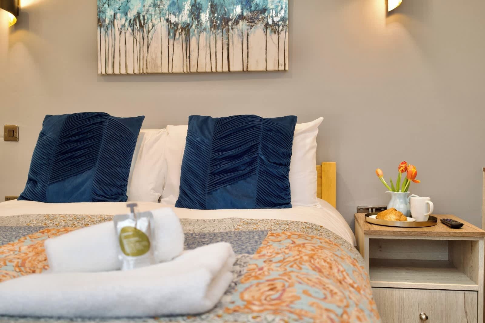 A neatly made bed with a patterned bedspread, two pillows, a bedside table with a vase of flowers, and a tray with a croissant and a mug.