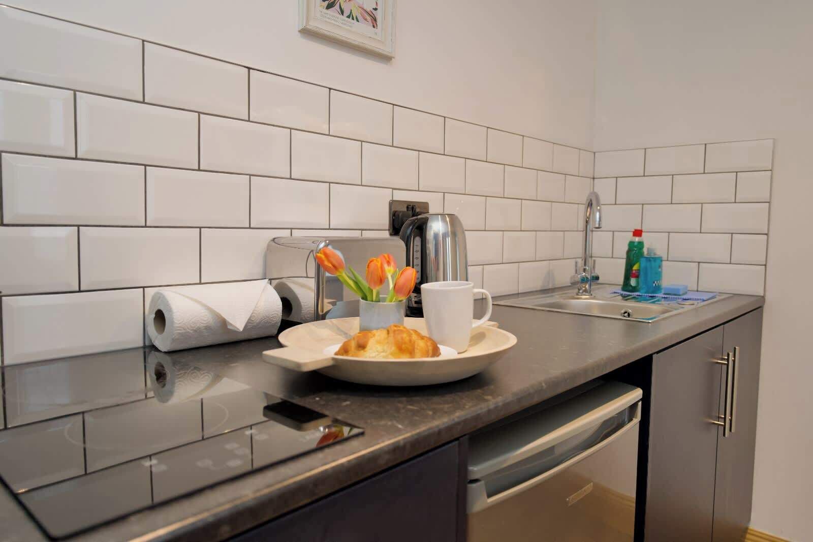 A kitchen with white tiled backsplash, a coffee maker, a roll of paper towels, a plate with a croissant and a cup, and a sink with cleaning supplies.