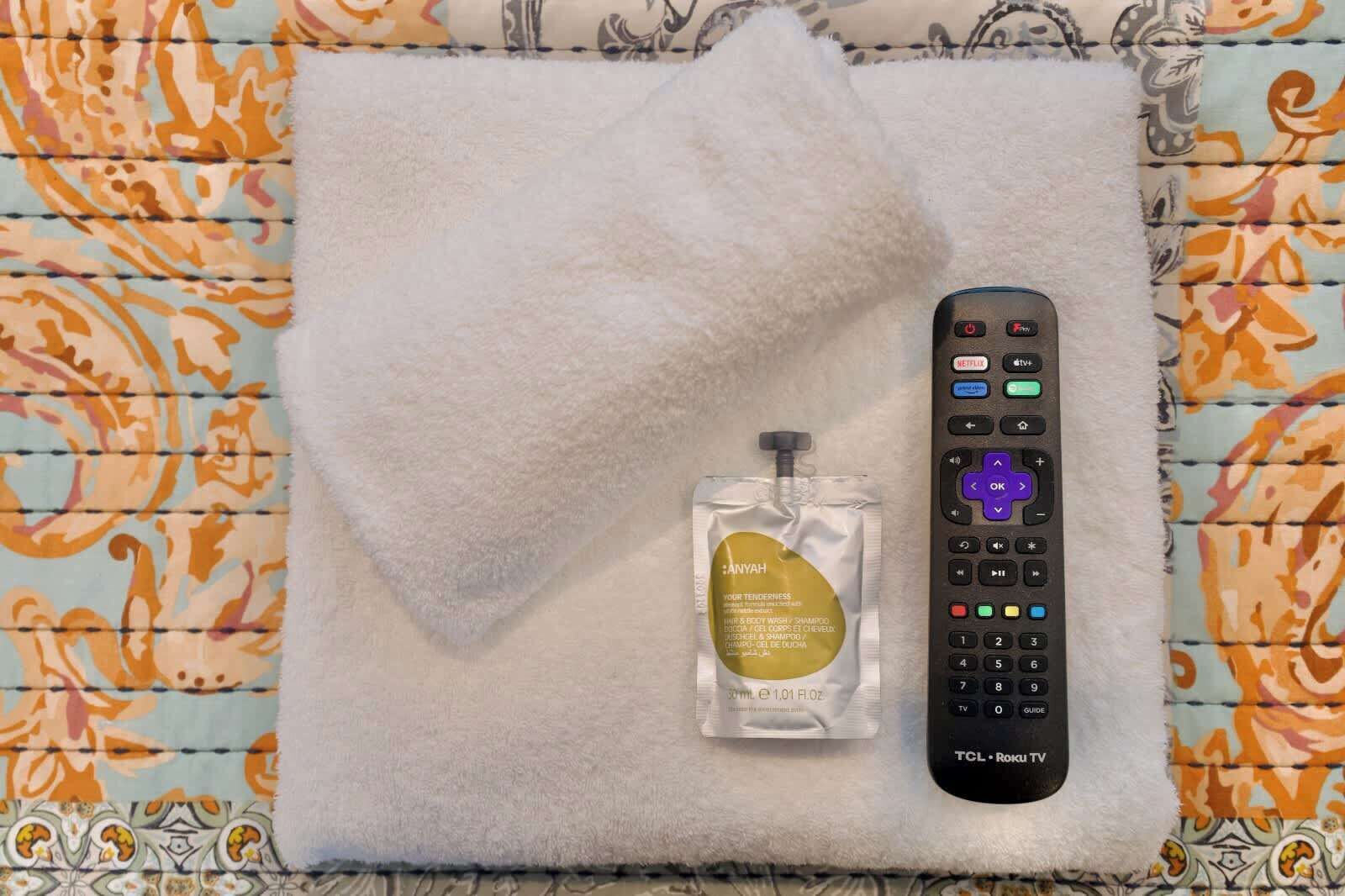 A remote control, a bottle of shampoo, and a white towel on a patterned fabric surface.