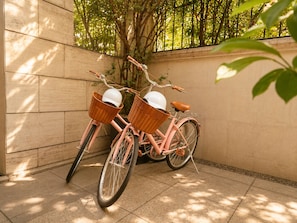 Una bicicleta rosa con dos cestas de mimbre estacionada frente a una pared con un árbol y una cerca al fondo.