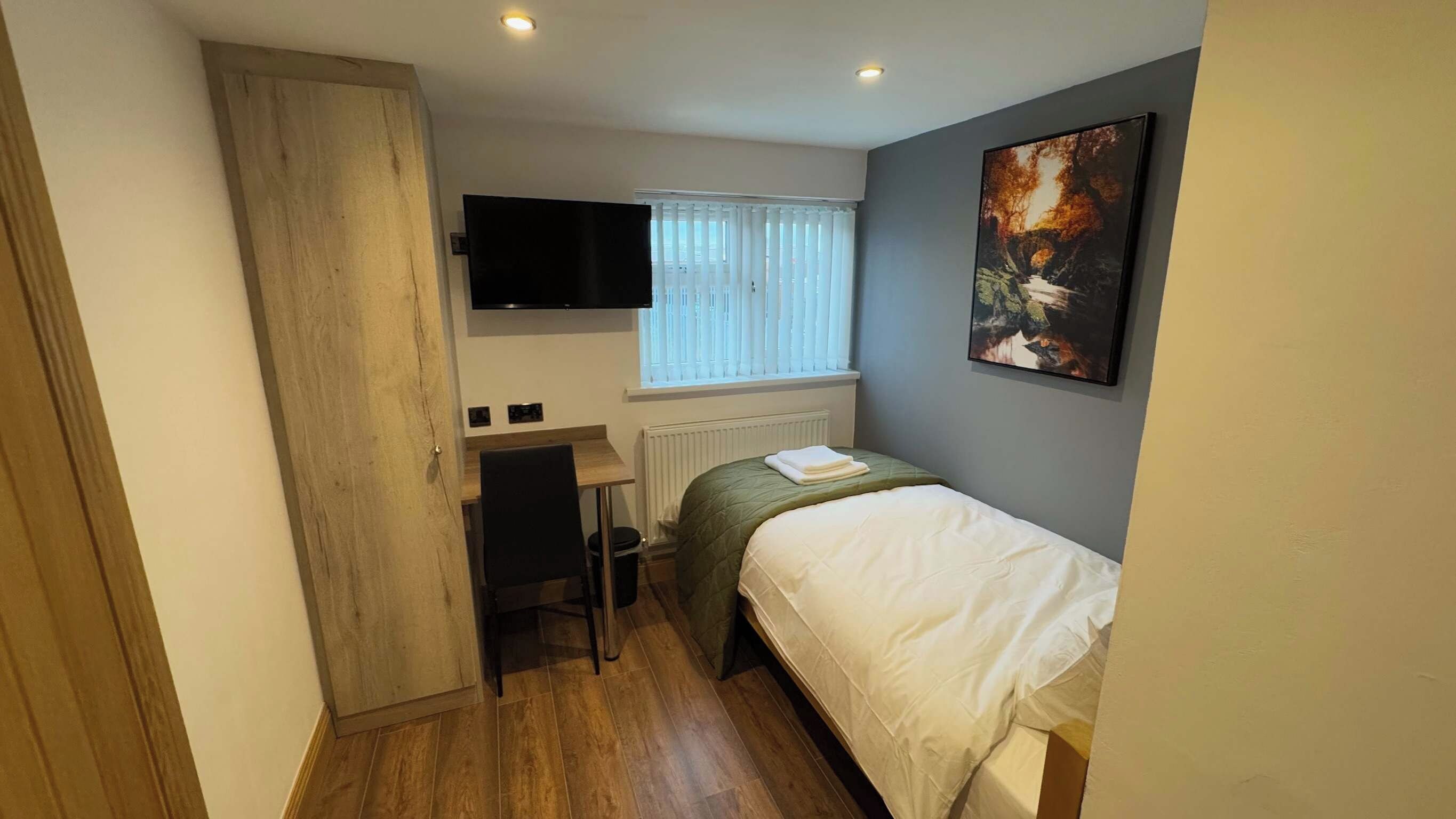 A small, well-lit bedroom with a bed, a desk, a chair, a radiator, and a TV mounted on the wall.