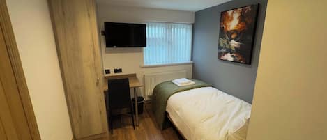 A small, well-lit bedroom with a bed, a desk, a chair, a radiator, and a TV mounted on the wall.