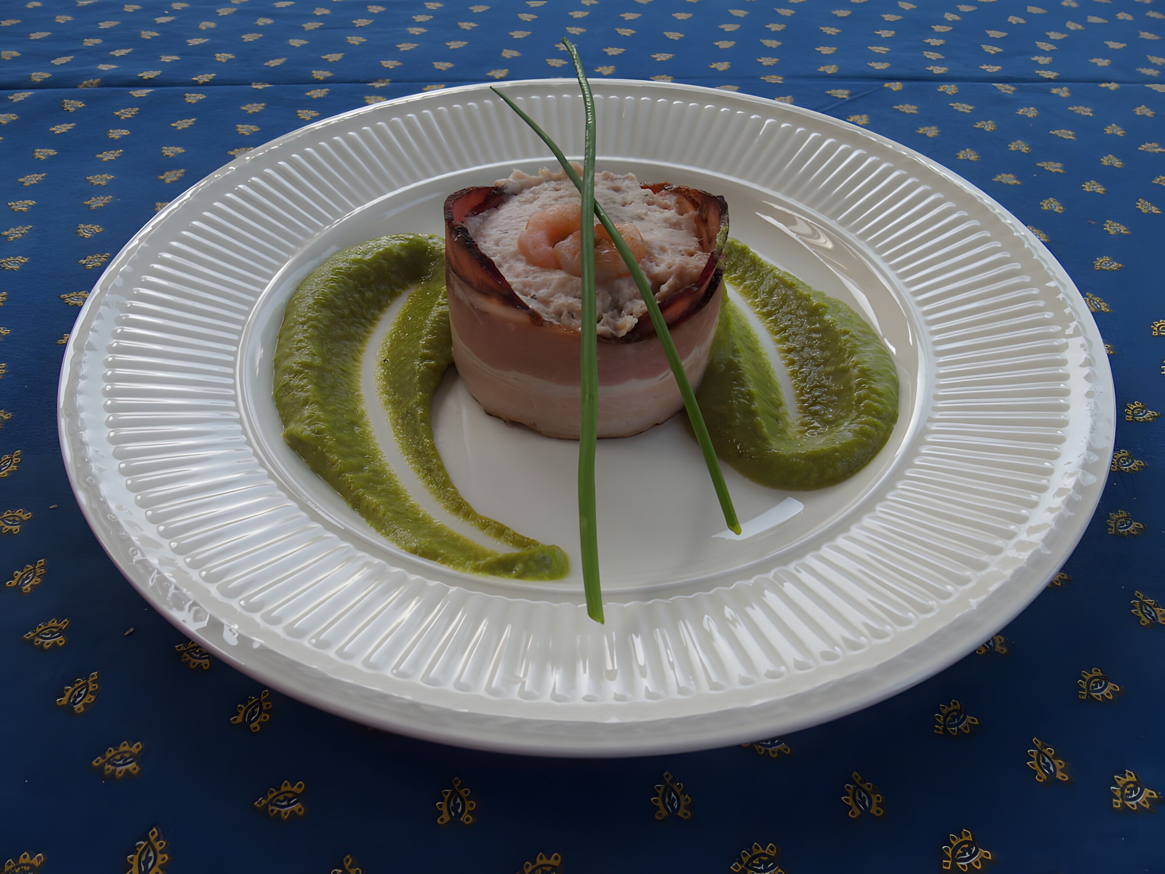 Un plato blanco con un alimento en el centro, adornado con salsa verde y un cebollino, sobre un mantel azul estampado.