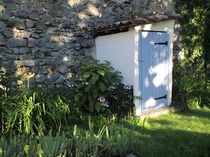 A stone wall with a small shed, surrounded by lush greenery and flowering plants. - Holiday Home 'Gîte La Cerisaie' with Mountain Views, Shared Pool, and Wi-Fi (Saint-Prix)