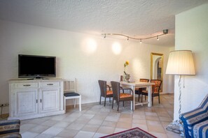 A living room with a television, dining table, chairs, and a lamp. - Apartment '104 Arnika' with Mountain View, Balcony and Wi-Fi (Riezlern)
