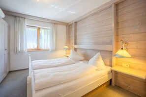 A double bed with white linens, a wooden headboard, two bedside lamps, a window with curtains, and a carpeted floor. - Apartment '104 Arnika' with Mountain View, Balcony and Wi-Fi (Riezlern)