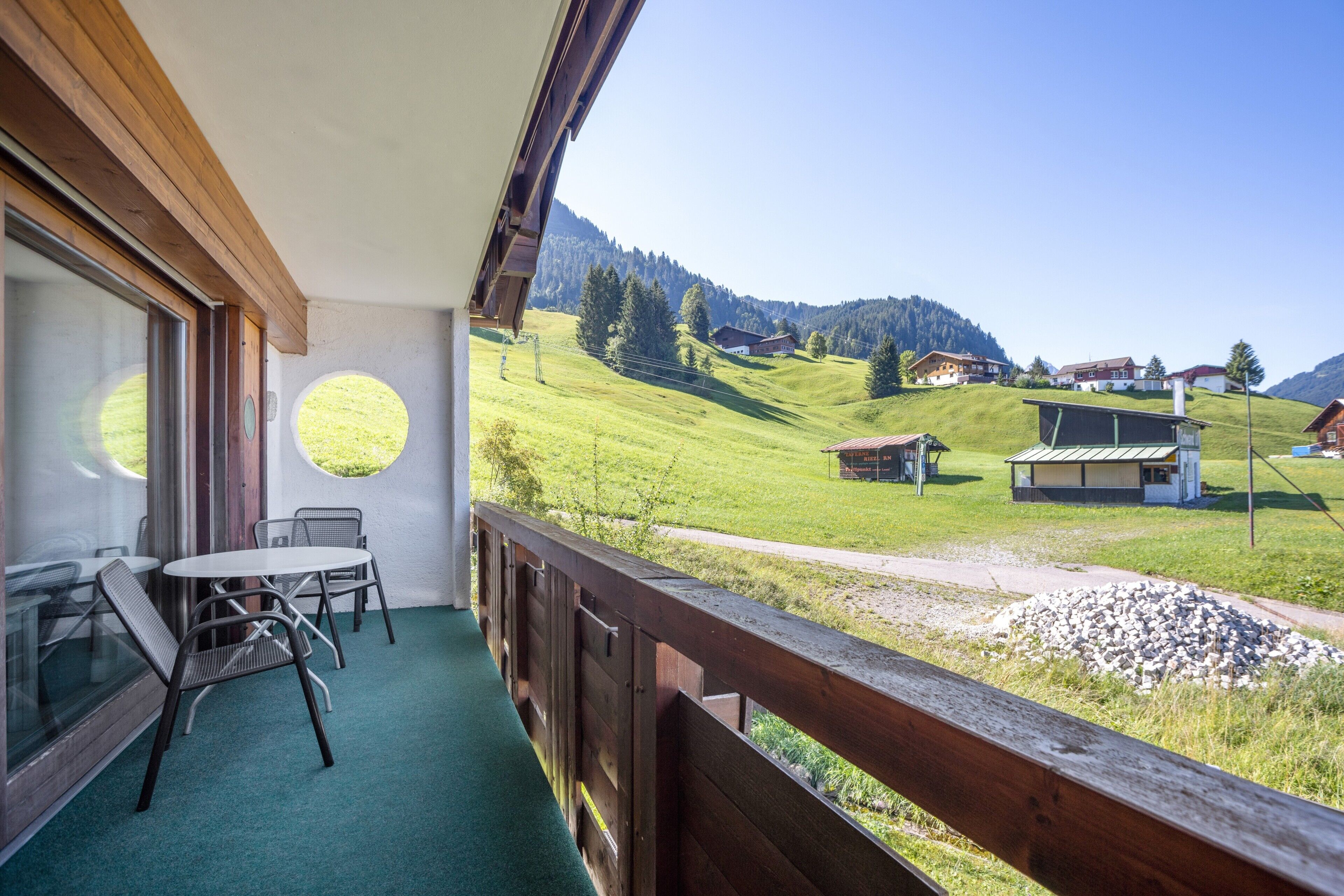 Un balcon donnant sur une vallée verdoyante et sur des maisons, une table avec des chaises et une balustrade en bois.