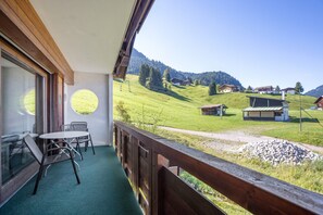 A balcony with a view of a green valley and houses, a table with chairs, and a wooden railing. - Apartment '104 Arnika' with Mountain View, Balcony and Wi-Fi (Riezlern)