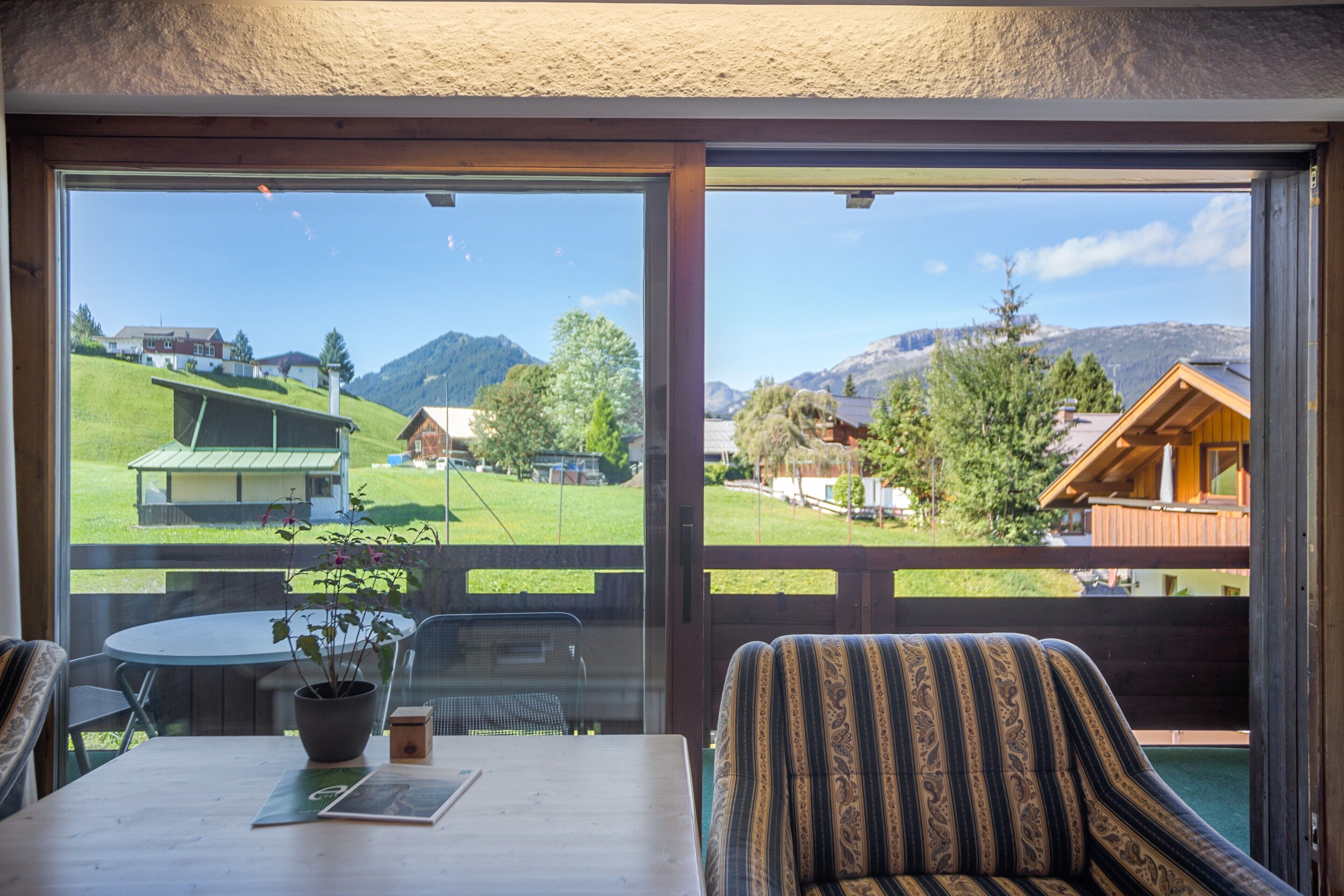 Een kamer met een groot raam met uitzicht op een groen landschap en bergen. Er staat een tafel met een plant en een stoel met een gestreept kussen.