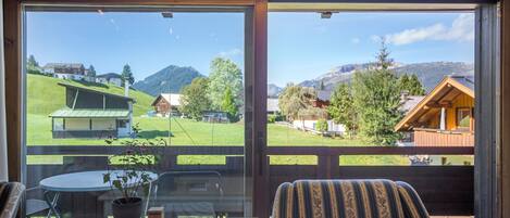 A room with a large window offering a view of a green landscape and mountains. There is a table with a plant and a chair with a patterned cushion.
