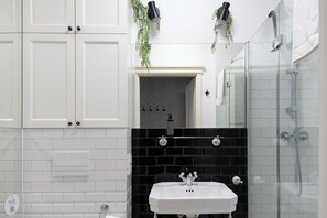 Casa de banho com armários brancos, painel de azulejos pretos, uma pia branca e cabine de chuveiro com divisória de vidro.