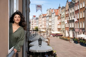 A woman standing by a window overlooking a street with buildings and outdoor seating. - Neptun View - Gdańsk Old Town  (Gdańsk)