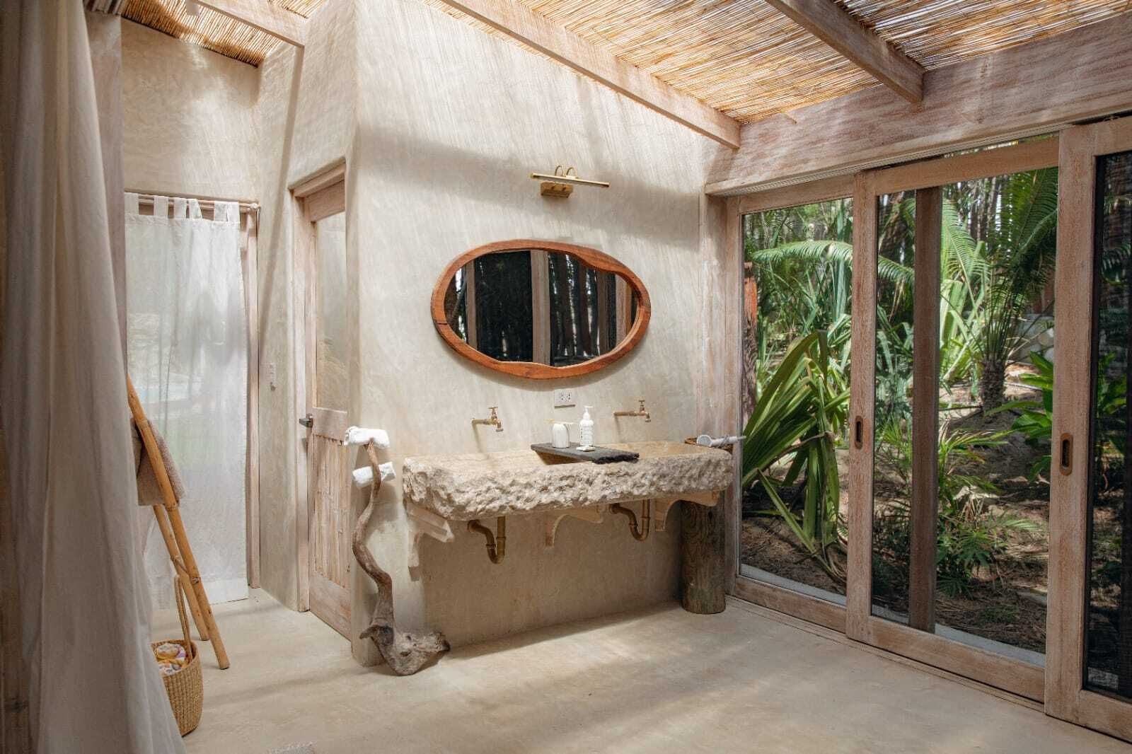 Une salle de bain avec un lavabo en pierre, un miroir encadré en bois et une vue sur un jardin tropical, visible à travers une porte vitrée.