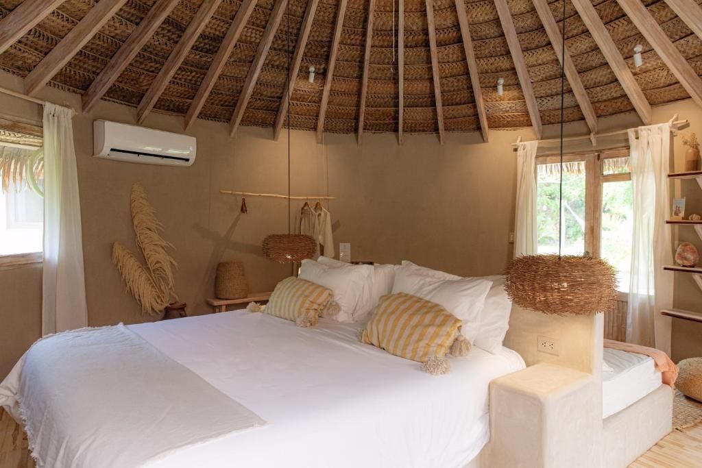 Une chambre à coucher avec un toit de chaume, un lit recouvert de draps blancs, une table de chevet et une étagère ornée d’objets décoratifs.