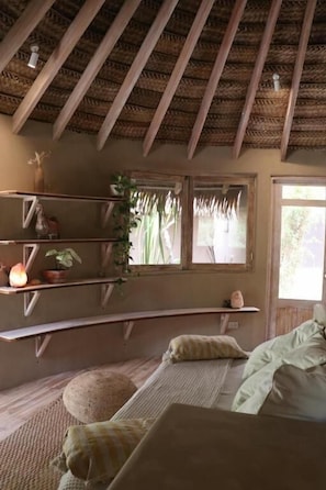 Una habitación con un banco de madera curvo, una cama, una estantería con plantas y un ventanal con cortinas.