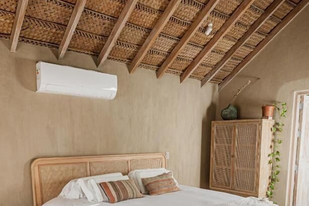 Un dormitorio con cama de madera, cabecera tejida, armario de fibra natural y una planta en maceta.