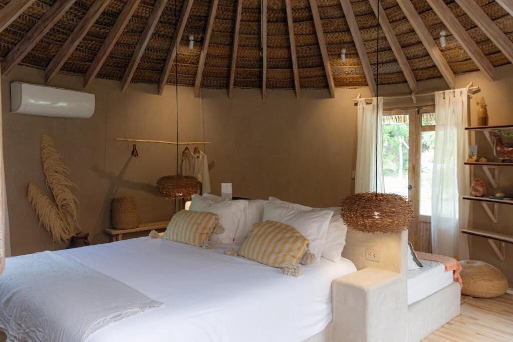 Un dormitorio con cama, almohadas, una lámpara colgante y un ventanal con cortinas.