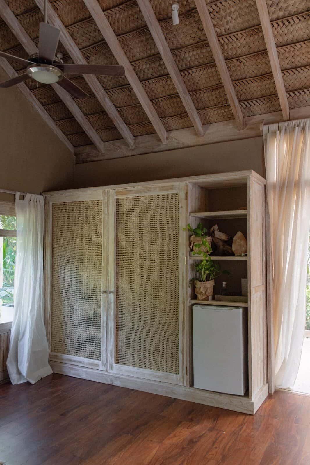 A room with a ceiling fan, a woven ceiling, a cabinet with shelves, and a small refrigerator.