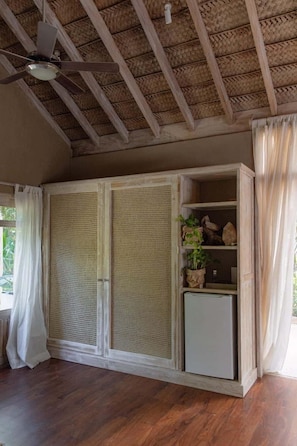 Una habitación con ventilador de techo, techo tejido, un mueble con repisas y un refrigerador pequeño.