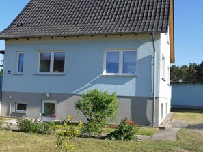 A two-story residential building with a grey tiled roof, white walls, and a small front garden with various plants. - Feldmann Holiday Accommodations (Korswandt)