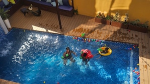 Une piscine avec une terrasse en bois, un coin salon doté de coussins et des plantes en pot.