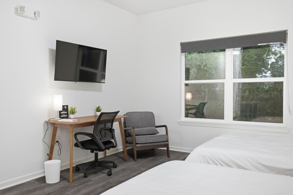 A room with a bed, a desk with a chair, a television mounted on the wall, and a window with a view of trees.
