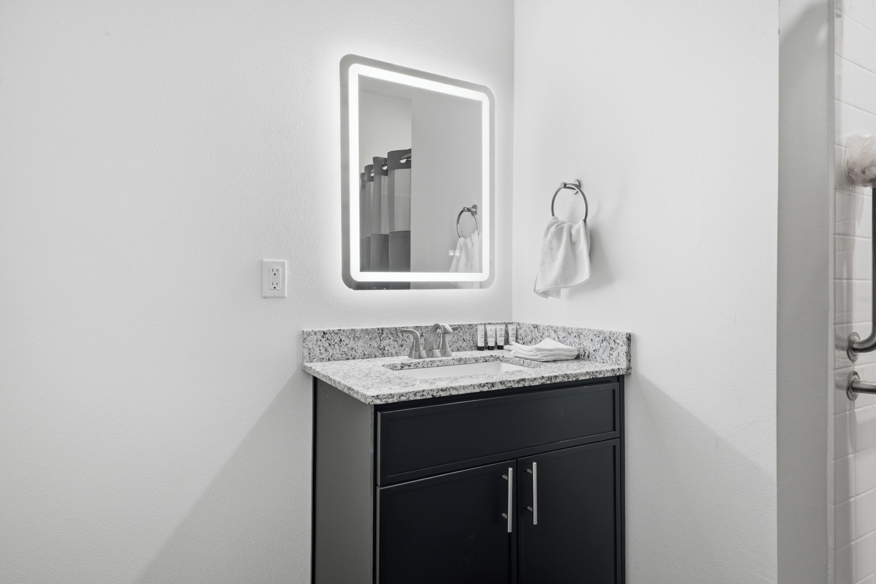 A modern bathroom with a granite countertop, a black cabinet, a wall-mounted mirror, and a towel rack.