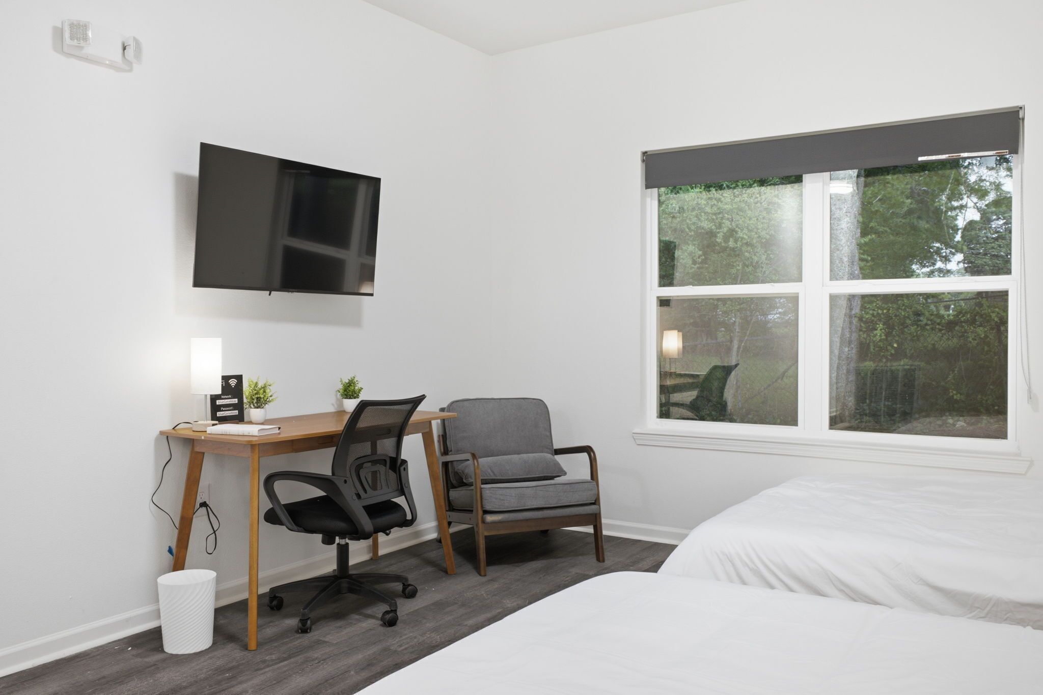 Een slaapkamer met een bed, een bureau met stoel, een aan de muur bevestigde televisie en een raam met uitzicht op bomen.
