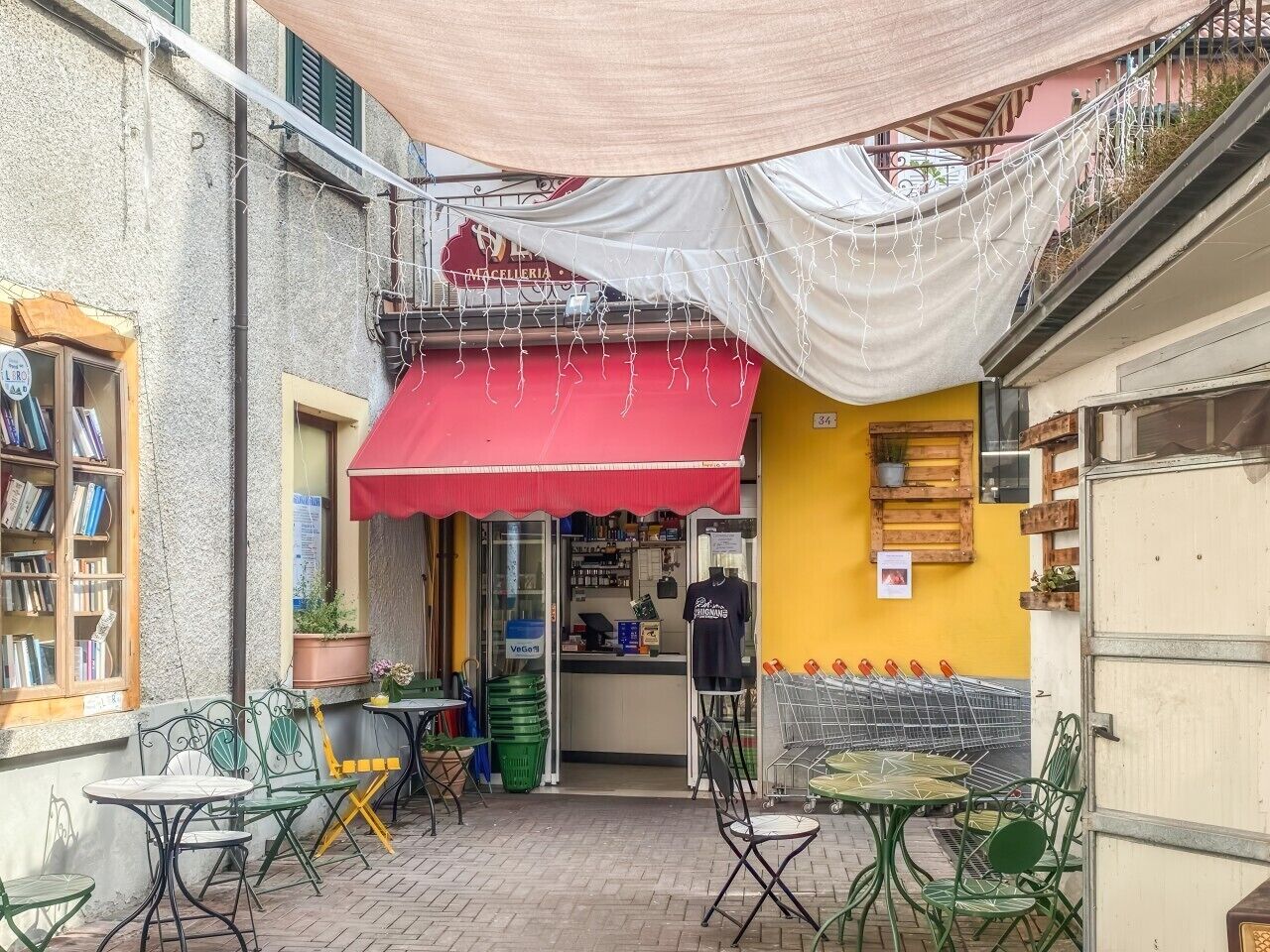An outdoor café with tables and chairs, a red awning, and a yellow wall with a wooden shelf.