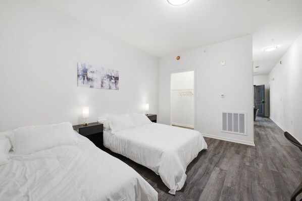 A hotel room with two single beds, a bedside table with a lamp, a wall-mounted artwork, a door leading to a bathroom, and a ventilation system on the wall.