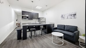 Una cocina moderna con armarios oscuros, una isla central con taburetes y un sofá gris en la sala.