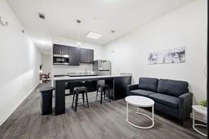 A modern kitchen with dark cabinets, a central island with stools, and a grey sofa in a living area.