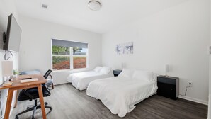 Un dormitorio moderno con dos camas individuales, un escritorio con silla, una ventana con vista a follaje y un cuadro en la pared.