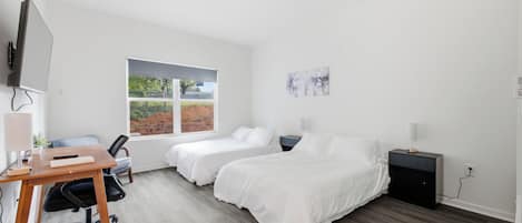 Un dormitorio moderno con dos camas individuales, un escritorio con silla, una ventana con vista a follaje y un cuadro en la pared.
