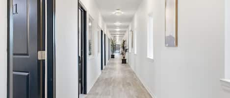 A long, narrow hallway with a light-colored wooden floor, white walls, and a series of doors on one side.