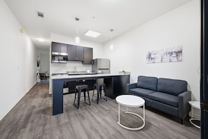 A modern kitchen with dark cabinets, a central island, and bar stools. There is a gray sofa and a small white table in the living area.