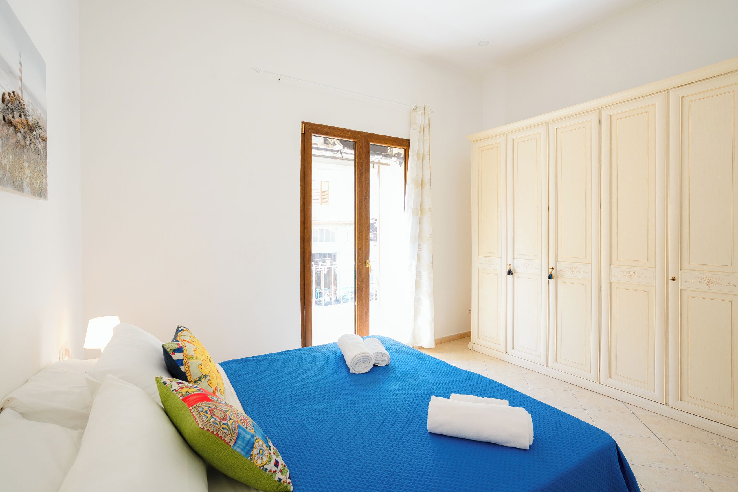 A bedroom with a bed, pillows, a blue bedspread, a wardrobe, and a door leading outside.