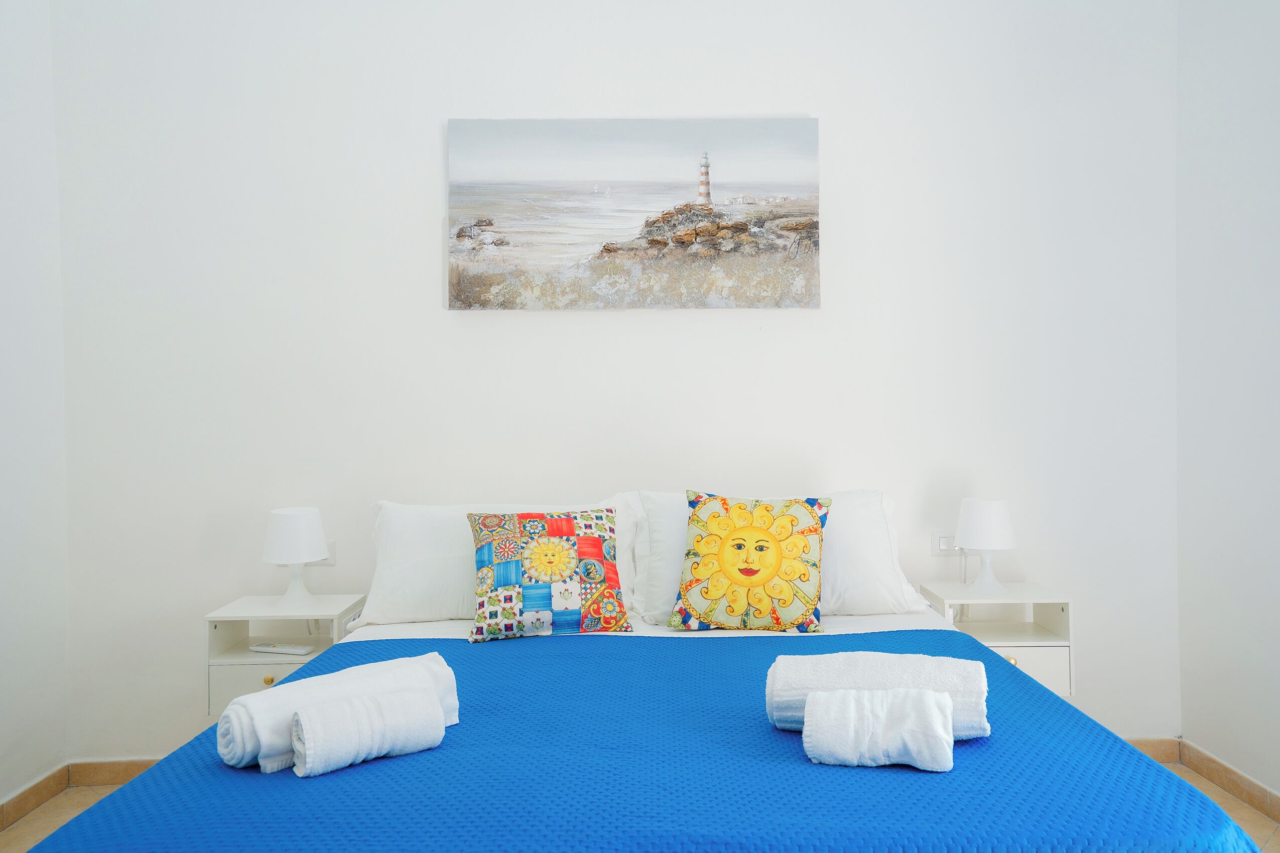 A hotel room with a bed, two pillows, a painting of a lighthouse, and two bedside tables with lamps.
