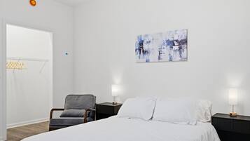 Un dormitorio con cama, dos mesitas de noche con lámparas, una silla y una obra de arte colgada en la pared.