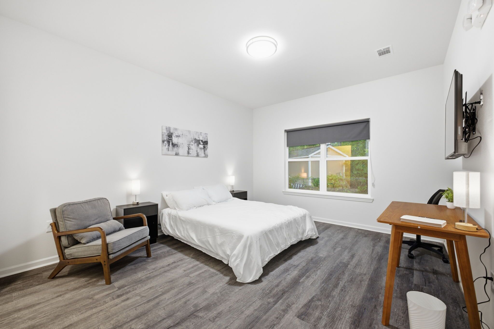 A bedroom with a bed, a chair, a desk, a window, and a wall-mounted TV.