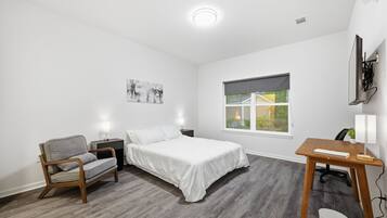 A bedroom with a bed, a chair, a desk, a window, and a wall-mounted TV.