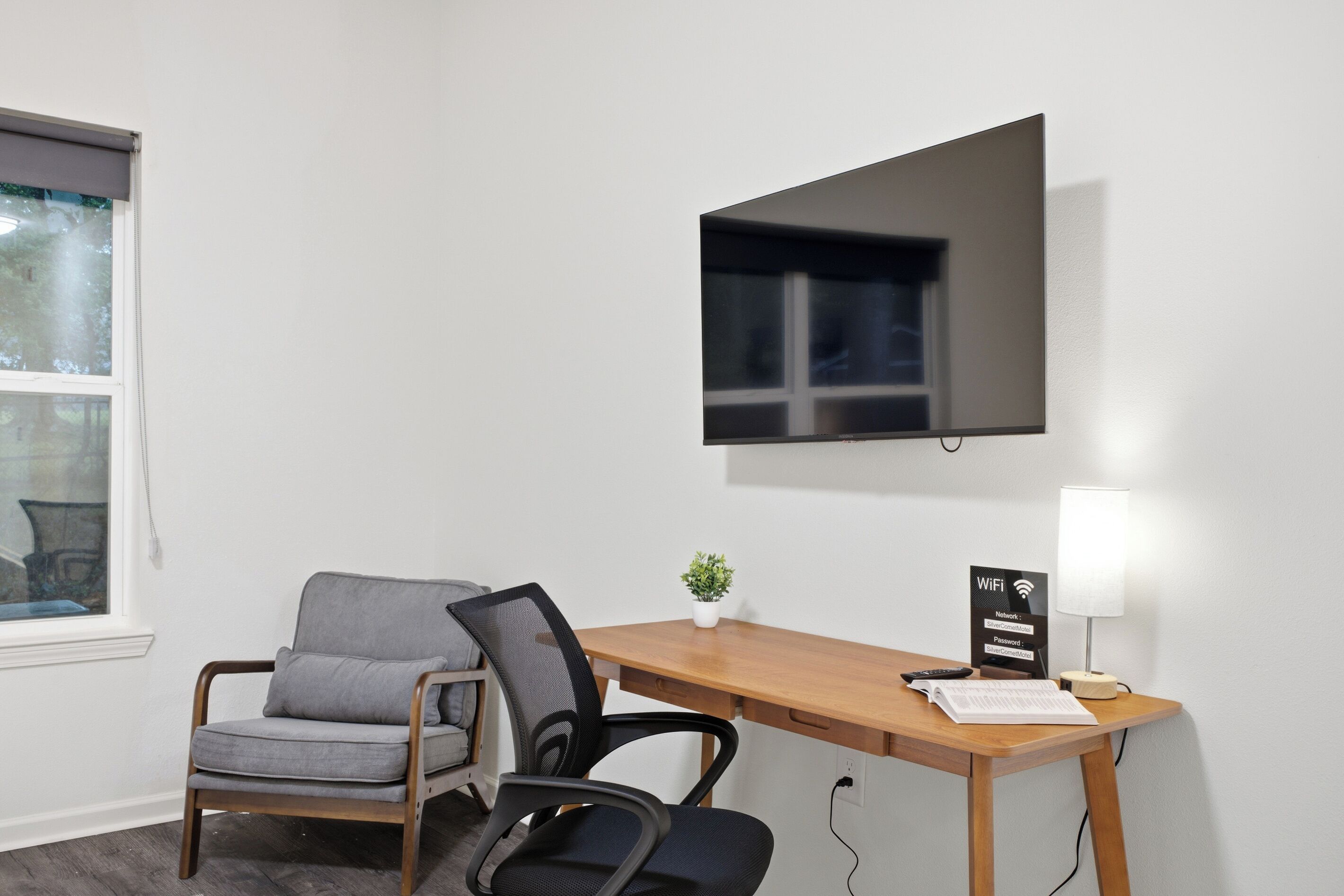 Habitación de hotel con escritorio de madera, una silla, televisor montado en la pared, una planta pequeña sobre el escritorio y un cartel de Wi-Fi.