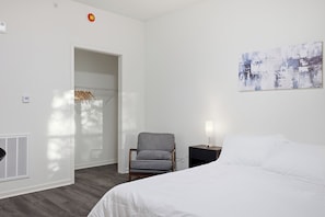 A hotel room with a bed, a chair, a nightstand, a painting, and a closet with clothes.
