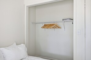 Un dormitori amb un llit, un armari amb una barra de fusta per a penjadors i una paret blanca.