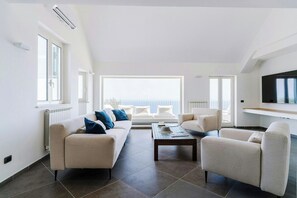 A modern living room with a sofa, armchairs, and a coffee table. There is a TV mounted on the wall and a window providing a view of the outdoors. - Villa near Monte San Costanzo with pool (Massa Lubrense)
